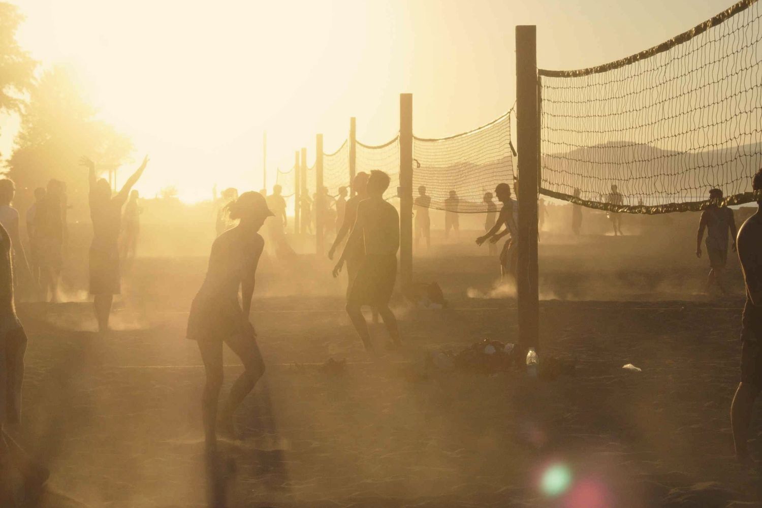 Backlit beach volleyball games at sunset, players in dust and lens flare.