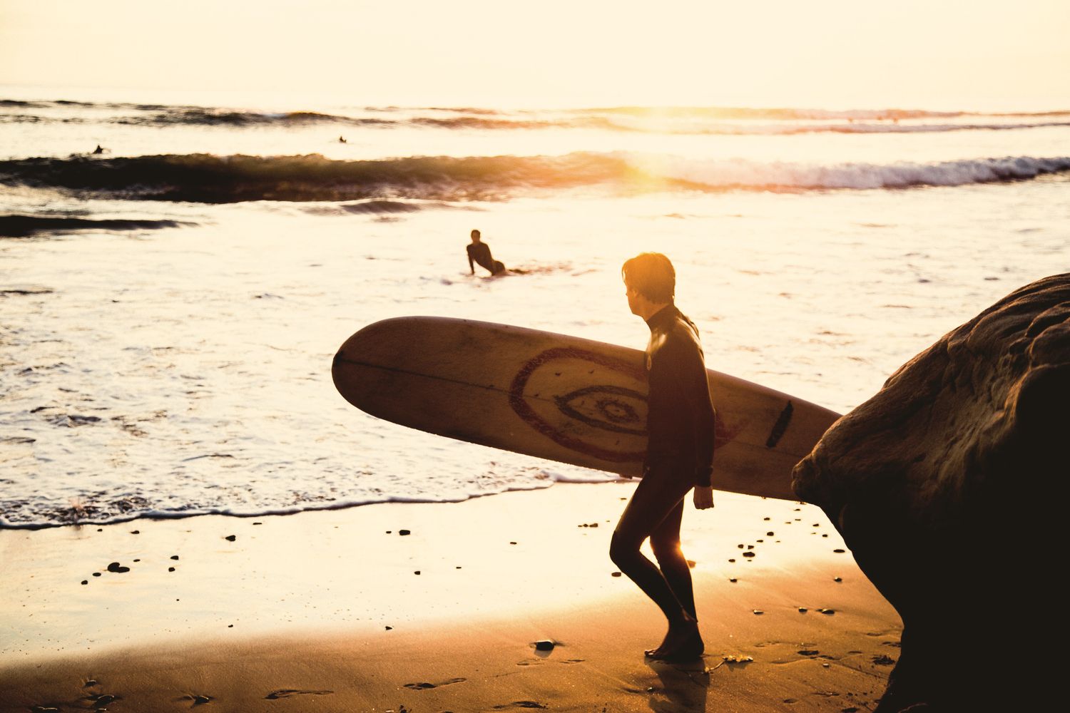 A surfer walks along the shoreline carrying a longboard, backlit by a low sun.